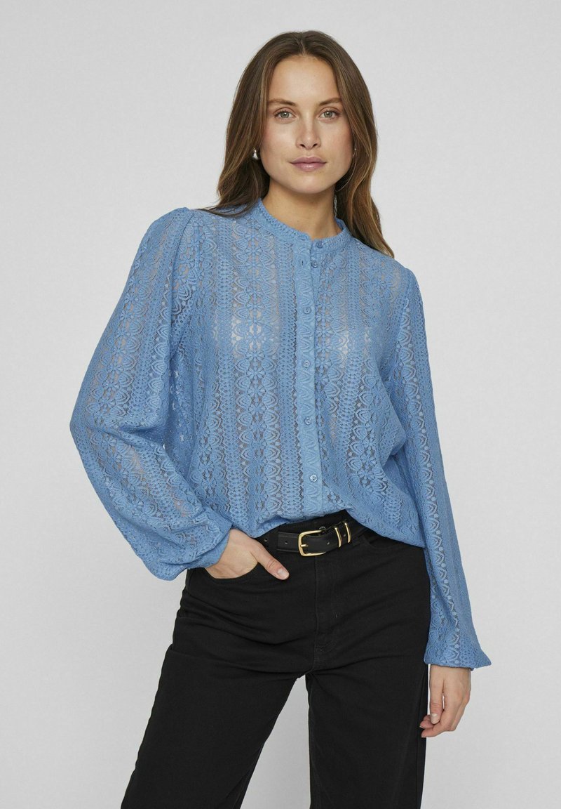 Blouse en dentelle bleu clair avec un col rond, des boutons à l'avant et des manches longues. Présente des motifs délicats et une coupe décontractée. Associée à un pantalon noir.