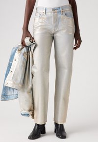 Jeans in argento metallico a vita alta con taglio dritto, abbinati a una giacca di jeans azzurro chiaro. La modella indossa stivali neri con tacco grossso.