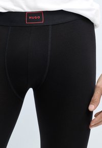 Černé elastické legíny s těsným střihem, vybavené širokým elastickým pasem a červenou štítkovou nálepkou s nápisem "HUGO". Hladký povrch a žádné vzory.
