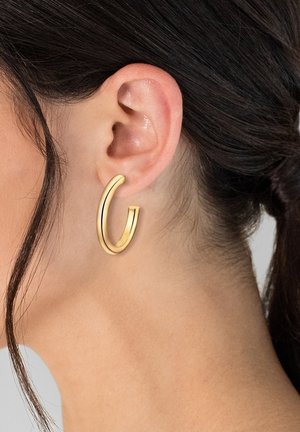 Goldene Creolen mit einer gebogenen Form, glatter Textur und polierter Oberfläche, getragen an einem Ohr mit nach hinten gestyltem dunklem Haar.