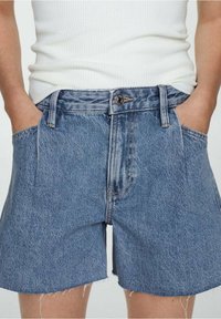 Persona que lleva pantalones cortos de mezclilla azul con un dobladillo sin terminar y las manos en los bolsillos, combinados con una parte superior sin mangas de canalé blanca.