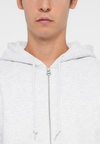 Gray zip-up hoodie with a drawstring hood, made of soft material featuring subtle texture, and a small brand logo on the chest.