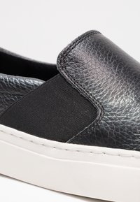 Chaussure à enfiler en cuir noir texturé avec empiècement élastique sur le côté et semelle en caoutchouc blanche, présentée sur un fond clair uni.