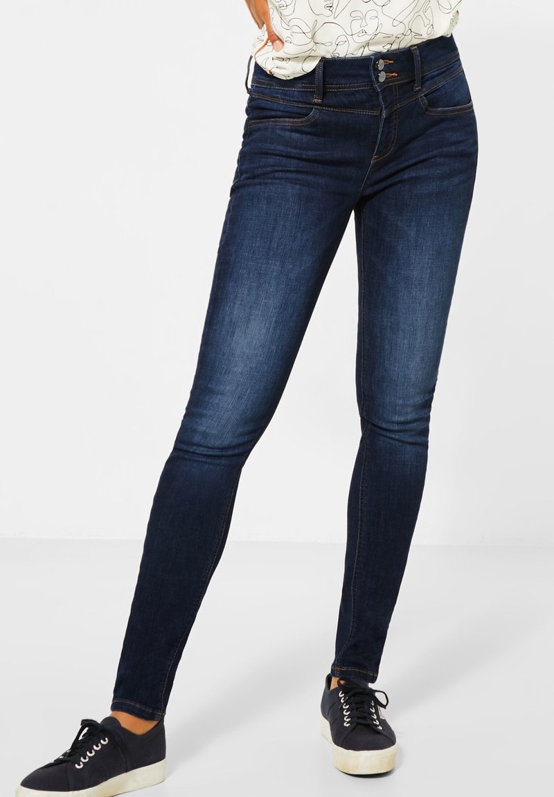 Femme portant un jean skinny bleu foncé, des baskets noires à lacets avec semelles blanches, et une chemise blanche avec un motif abstrait de visage en ligne.
