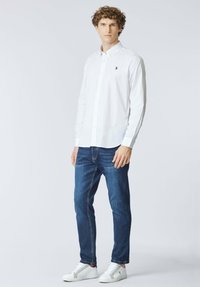 Camicia bianca con bottoni e maniche lunghe, logo ricamato blu sul petto sinistro, abbinata a jeans blu scuro e sneakers bianche.