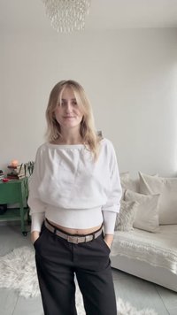 Λευκό, υπερμεγέθες πουλόβερ με φαρδύ ντεκολτέ, σε συνδυασμό με ψηλόμεσο μαύρο παντελόνι και ανοιχτό καφέ ζώνη. Φωτεινή διακόσμηση δωματίου στο παρασκήνιο.