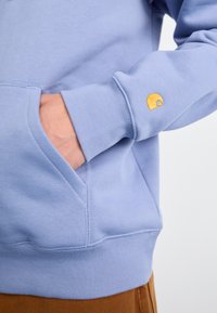 Sweatshirt bleu clair avec des poignets et un ourlet côtelés, dotée d'une poche avant et d'un petit logo jaune sur la manche gauche. Texture douce.