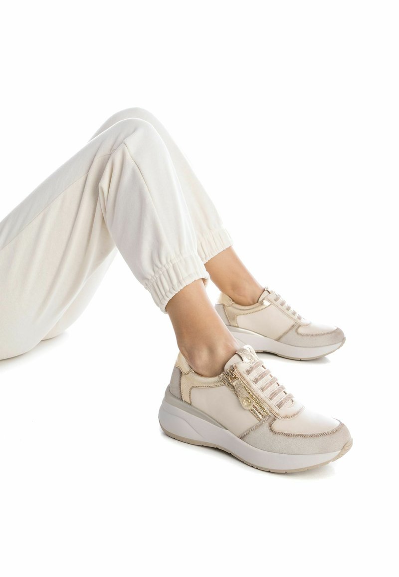 Zapatilla de color beige claro con una parte superior texturizada, cremallera lateral y cordones elásticos. El diseño incluye una suela gruesa y acolchada.