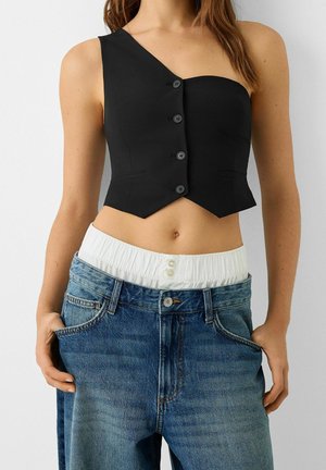 Kvinde iført asymmetrisk sort knappet crop top og løse blå jeans med hvid elastisk linning, hænderne i lommerne, foran hvid baggrund.