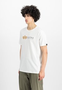 Balta medvilninė marškinėlių su apvaliu kaklu, su auksiniu "ALPHA INDUSTRIES" logotipu ir juodu etikete ant rankovės.