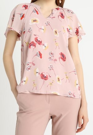 Femme portant un chemisier floral rose doux à manches volantées, associé à un pantalon rose clair assorti.