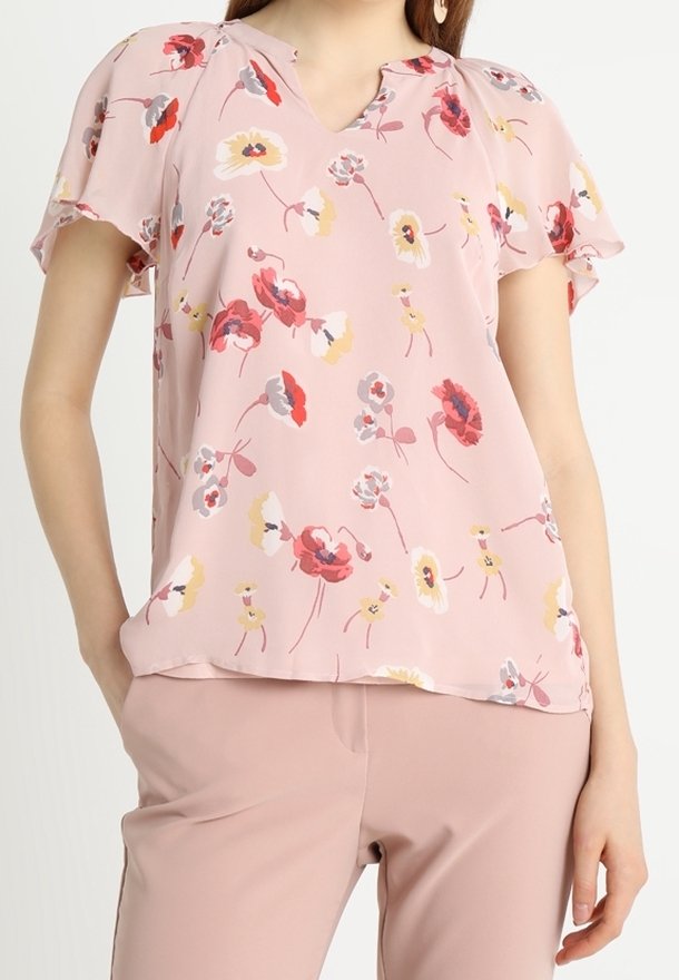 Femme portant un chemisier floral rose doux à manches volantées, associé à un pantalon rose clair assorti.