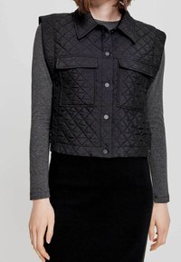 Czarna pikowana kamizelka w rombowy wzór, z kołnierzem, dwoma przednimi kieszeniami i zapięciem na guziki, noszona na szarym longsleeve.