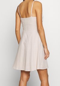 Kvinna som bär en ärmlös, ljusbeige, knälång klänning med tunna axelband och en dragkedja bak, stående mot en enkel bakgrund.