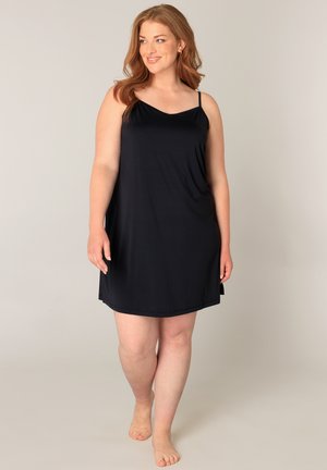 Vrouw met rood haar, gekleed in een zwarte mouwloze jurk, staat op blote voeten tegen een effen beige achtergrond en kijkt glimlachend naar links.