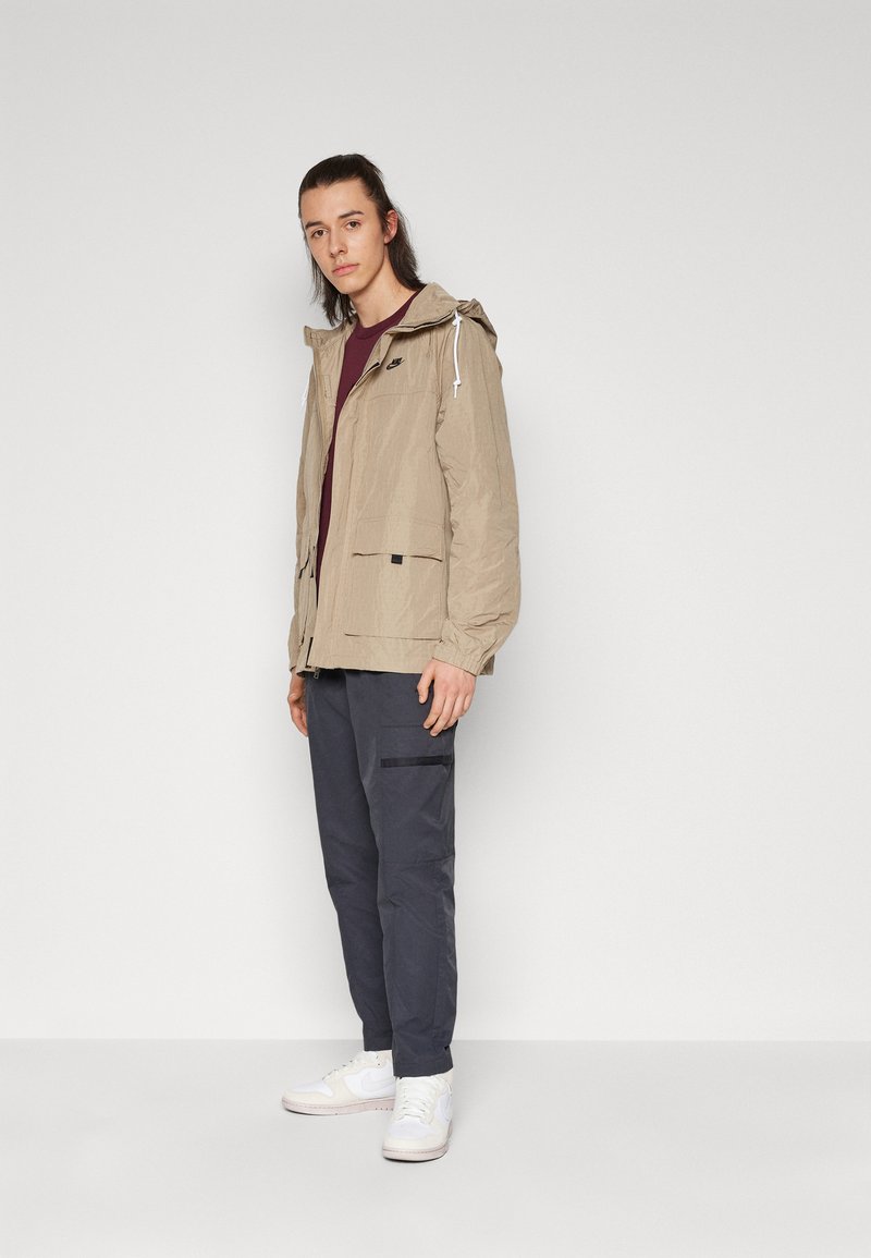 Chaqueta ligera beige con capucha ajustable, cierre con cremallera y bolsillos frontales, combinada con pantalones cargo oscuros y zapatillas blancas.