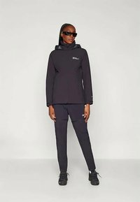 Donna che indossa una giacca e pantaloni neri da esterno con cappuccio e occhiali da sole, in piedi contro uno sfondo leggero e uniforme.