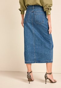 Falda midi de denim con silueta recta, color azul oscuro, dos bolsillos traseros y detalles de costura visibles. Combinada con tacones altos con estampado.