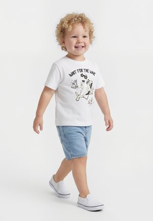 Niño con cabello rizado caminando, vestido con una camiseta blanca con un gráfico de tiburón y el texto "ESPERA LA OLA", shorts vaqueros y zapatos blancos sin cordones.