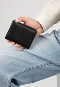 Portefeuille en cuir noir avec une surface texturée, fermeture éclair et forme rectangulaire compacte, tenu dans une main contre un denim bleu clair.