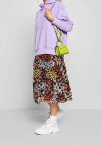 Sudadera púrpura clara con un bolsillo frontal, falda negra con flores y patrones en naranja, amarillo y blanco, bolso cruzado verde y zapatillas blancas.