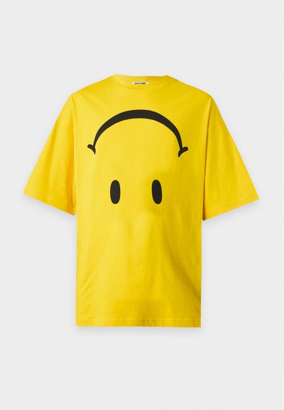 Κίτρινο T-shirt με κοντά μανίκια και μαύρο σχέδιο ακουστικών και δύο οβάλ σχήματα που μοιάζουν με μάτια στο μπροστινό μέρος.