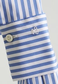 Tissu en coton rayé bleu clair et blanc, avec un bouton et un accent brodé. Poignet avec des détails de couture contrastants.