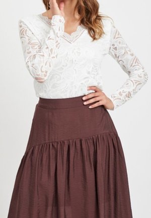 Femme portant un haut en dentelle blanche à manches longues et une jupe marron taille haute avec des détails froncés, main sur la hanche et touchant le visage.