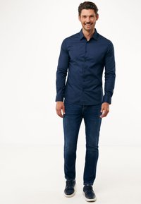 Camicia a maniche lunghe color blu navy, taglio slim, in cotone, abbinata a jeans scuri e sneakers blu navy, esposta su uno sfondo bianco.