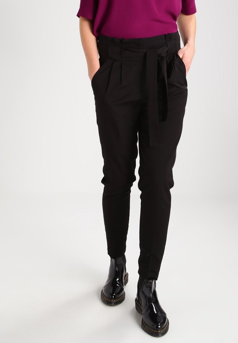 Pantalones ajustados negros con cinturón, que presentan una textura suave y un frente plisado, combinados con botas de tobillo negras brillantes.