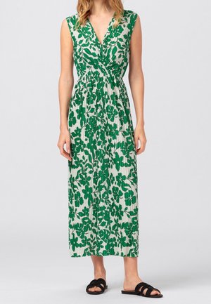 Ärmelloses weißes Maxikleid mit grünem Blumenmuster, plissiertem Stoff, V-Ausschnitt, tailliert, kombiniert mit schwarzen flachen Sandalen zum Hineinschlüpfen.