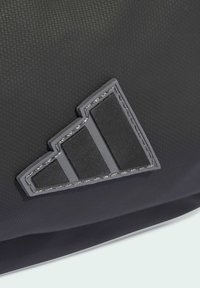 Material de mochila negra texturizada con un logo gris triangular que presenta tres rayas horizontales. Muestra costuras resistentes y un diseño minimalista.