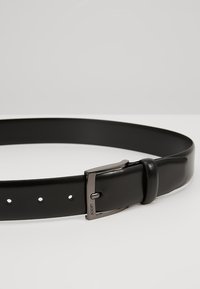 Svart lærbelte med en skinnende sølvspenne, en rekke perforeringer for størrelsejustering og en glatt, matt finish.