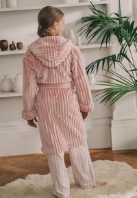 Roosa kapuutsiga hommikumantel tekstuurse triibulise kangaga, vööga taljes ja pehmete varrukate äärtega, kantud triibuliste pükstega pehmel, heledal vaibal.