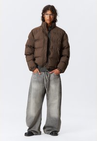 Brun pufferjakke med quiltet tekstur, glidelås og ribbestrikket kant, kombinert med løse lysegrå jeans og svarte sko.