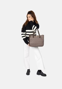 Brun mönstrad tote-väska med dubbla handtag, rektangulär form och guldemblem. Modellen bär en svart och vit randig tröja och vita byxor.