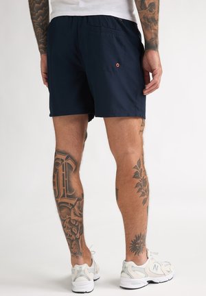 Marineblauwe zwembroek met elastische taille, achterzak en oranje oogje. Getoond wordt een gespierd been met ingewikkelde tatoeages.