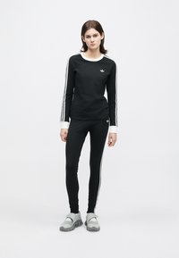 Mladý človek stojaci v čiernom Adidas teplákovom kompletne s bielymi pruhmi a šedých teniskách na hladkom bielom pozadí.