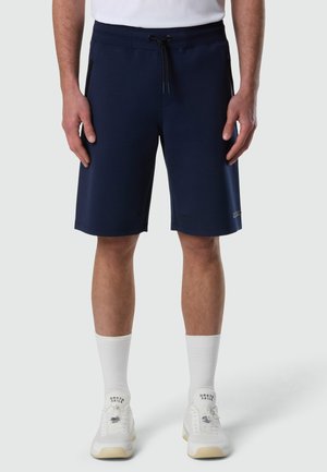 Uomo che indossa pantaloncini blu navy con coulisse, calzini bianchi e scarpe da ginnastica bianche, in piedi contro uno sfondo chiaro e uniforme.
