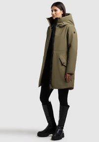 Parka impermeable verde oliva con un alto cuello, acentos negros, cierre frontal y bolsillos con botones a presión. Textura suave, hasta la rodilla, combinada con botas negras.