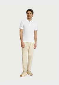 Valkoinen polo-paita lyhyillä hihoilla, teksturoitu kangas ja kolmen nappulan lista, yhdistettynä beigeihin housuihin ja vaaleisiin tennareihin.