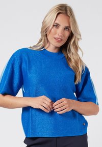 Top blu, a maniche corte in un tessuto a maglia luccicante. Presenta un collo rotondo e una leggera forma ad A. Dettaglio del logo dorato sull'orlo.