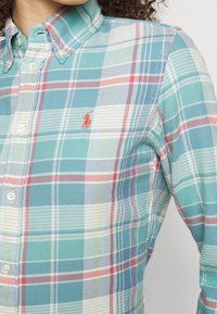 Zbliżenie na koszulę w kratę w kolorach morskim, czerwonym i białym z kołnierzykiem oraz małym haftowanym logo na piersi.