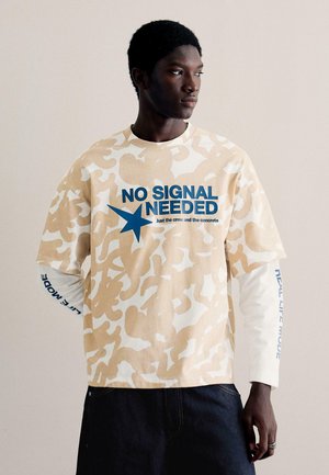 Czarnoskóry mężczyzna w beżowo-białej koszulce moro, nałożonej na białą koszulę z długim rękawem z niebieskim napisem, pozujący na tle o neutralnym kolorze.