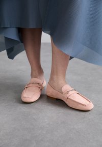 Pink suede loafers with a flat sole, featuring a decorative metal chain accent and stitch detailing. Worn with a pleated blue skirt.