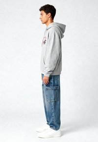 Felpa grigia con cappuccio e tasca frontale, caratterizzata da ricami di un cuore rosso e di un'ape. Indossata con jeans cargo blu larghi e sneakers bianche.