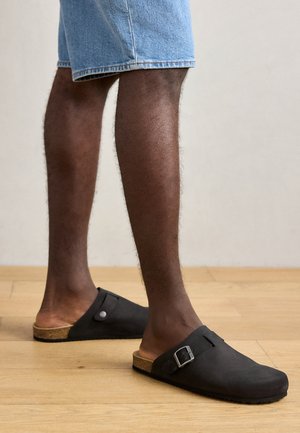 Zwarte instapklompen met een gespsluiting, kurken zool en zacht suede-achtig materiaal, gedragen met denim shorts op een houten vloer.