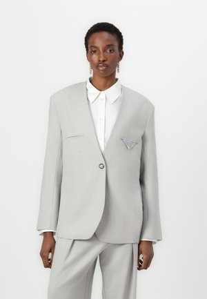 Blazer ajusté gris clair avec un design structuré, fermeture à un bouton, et un détail unique sur la poche poitrine. Porté sur une chemise blanche à col.