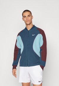 Chaqueta con cremallera de Nike que presenta una combinación de paneles en azul marino, azul claro y burdeos. Tejido suave con un ajuste ceñido y costuras en contraste.