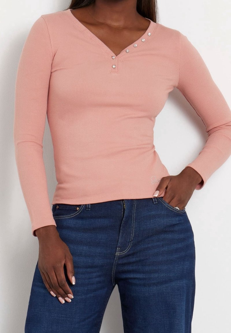 Haut à manches longues en maille côtelée de couleur rose doux, avec un décolleté en V orné de boutons. Porté avec un jean bleu foncé, présentant une texture lisse.